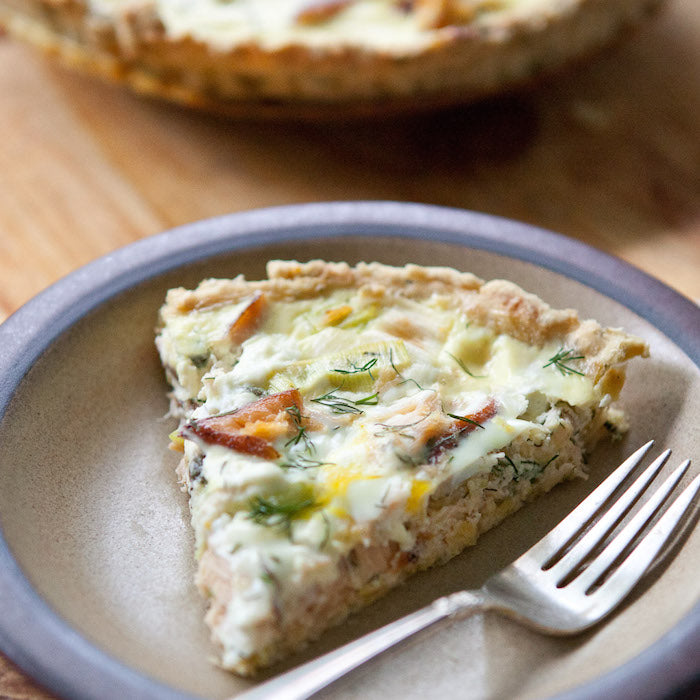 Close-up of a slice of smoked salmon tart with an Einkorn herbed crust, creamy filling and dill, on a ceramic plate with a fork