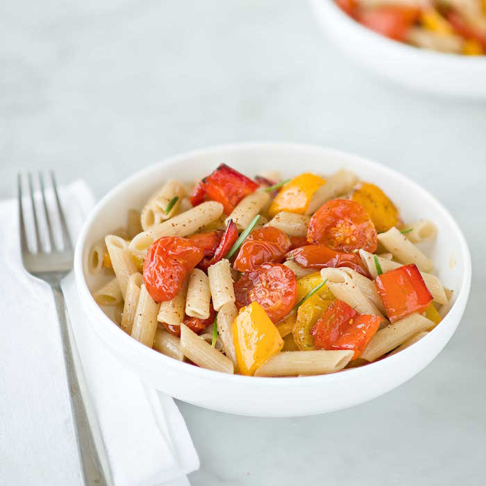 Bowl of Einkorn penne with oven-roasted cherry tomatoes, red and yellow bell peppers and rosemary, fork and napkin at side