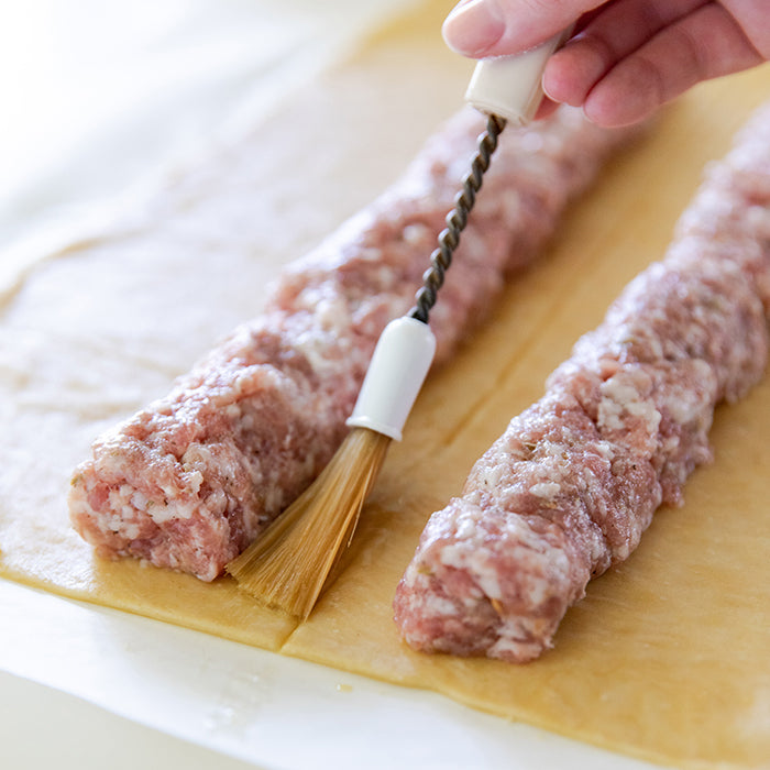 Two raw sausage logs on einkorn pastry being brushed with egg wash before rolling