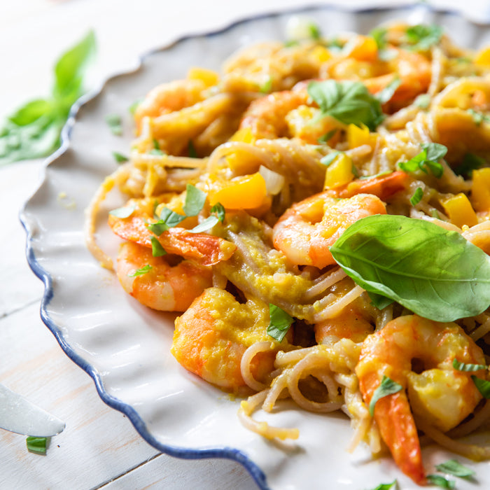Close-up of shrimp tossed with gluten-free cassava noodles, yellow pepper sauce, and fresh basil on a white scalloped plate