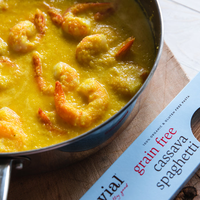 Shrimp simmering in yellow pepper sauce in a skillet beside a box of Jovial grain-free cassava spaghetti on a wooden board