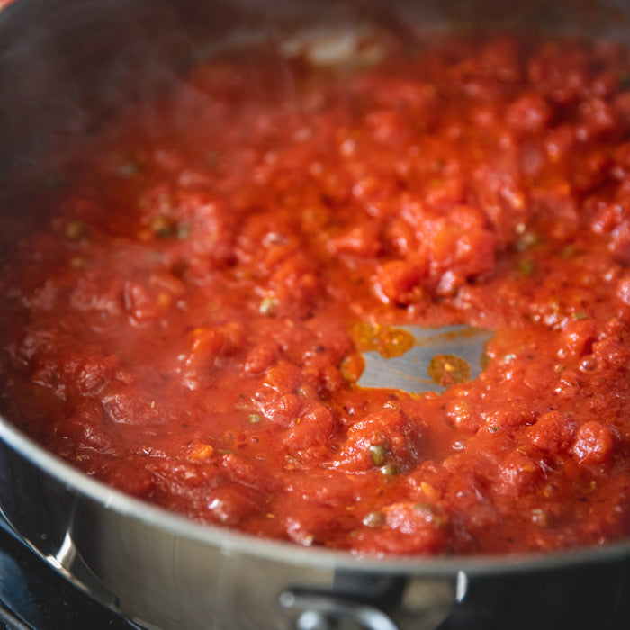 Puttanesca sauce simmering in a stainless-steel skillet with tomato chunks and capers