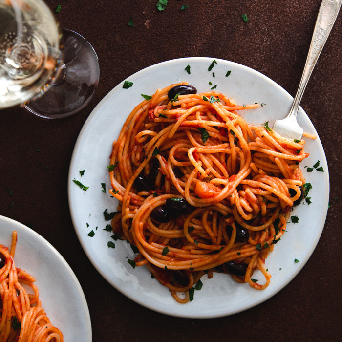 Plate of spaghetti alla puttanesca with black olives and parsley, fork on white plate beside a wine glass