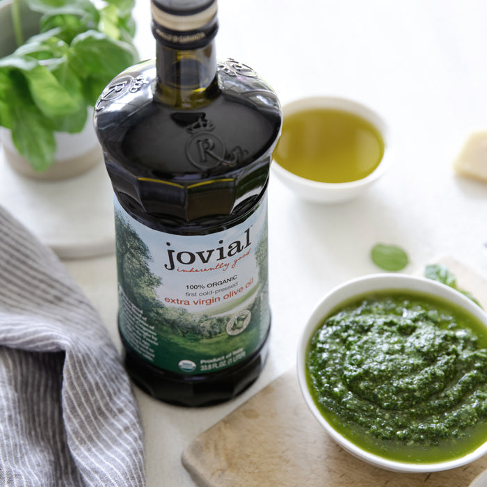 Jovial organic extra virgin olive oil bottle beside a bowl of fresh basil pesto and a small dish of oil on a light countertop