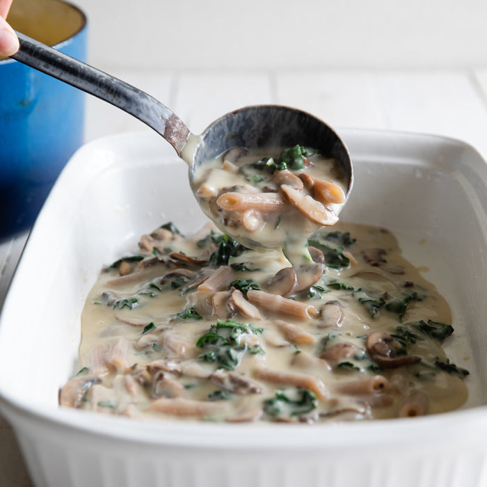 Ladle pouring baked penne in a creamy cauliflower sauce with mushrooms and kale into a white casserole dish