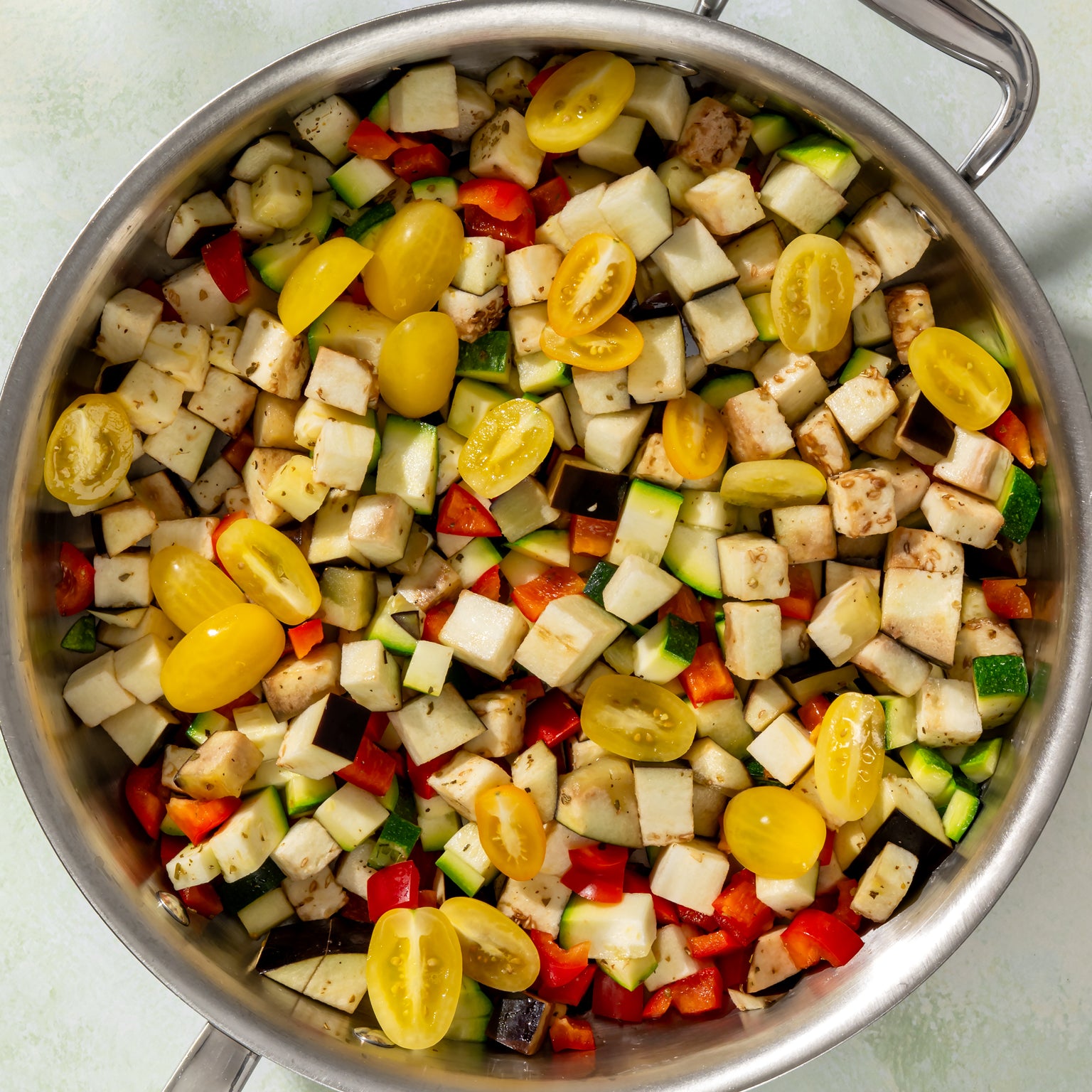 Diced zucchini, eggplant, red bell pepper, and yellow cherry tomatoes in a stainless skillet ready to cook