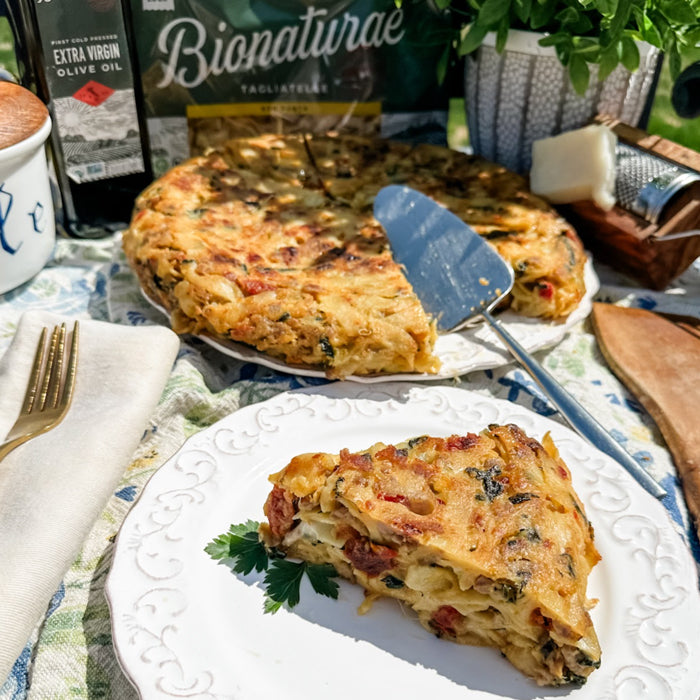 Close-up of a slice of sausage and spinach frittata with golden-brown crust and melted cheese, whole frittata and table setting in background
