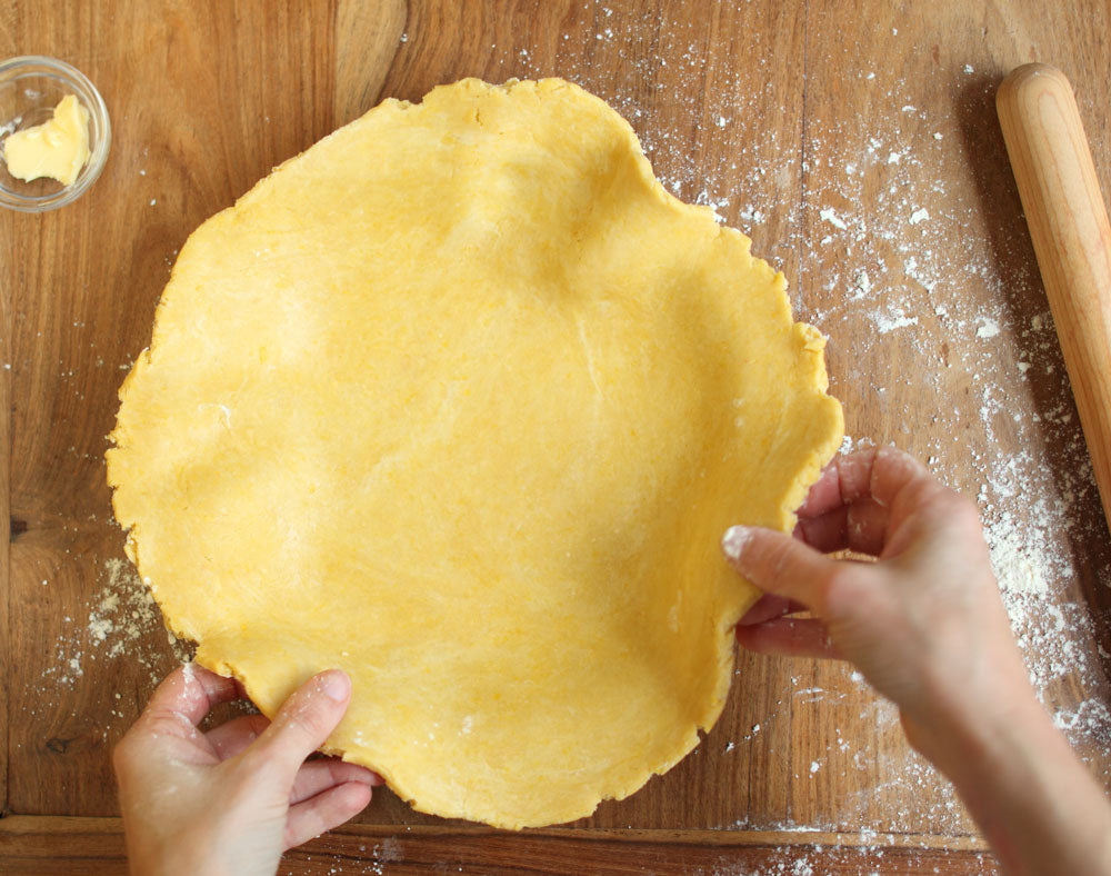Hands shaping an einkorn pie crust on a floured wooden board with a rolling pin and a small dish of butter nearby