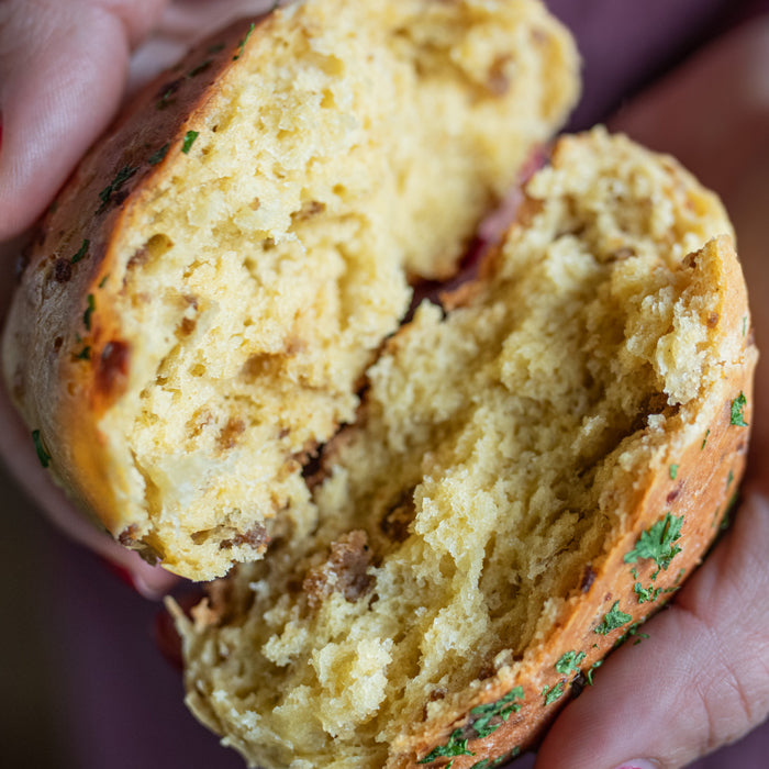 Close-up of split einkorn dinner roll showcasing sausage and melted cheese with a flaky interior and parsley-dusted golden crust, held in hands