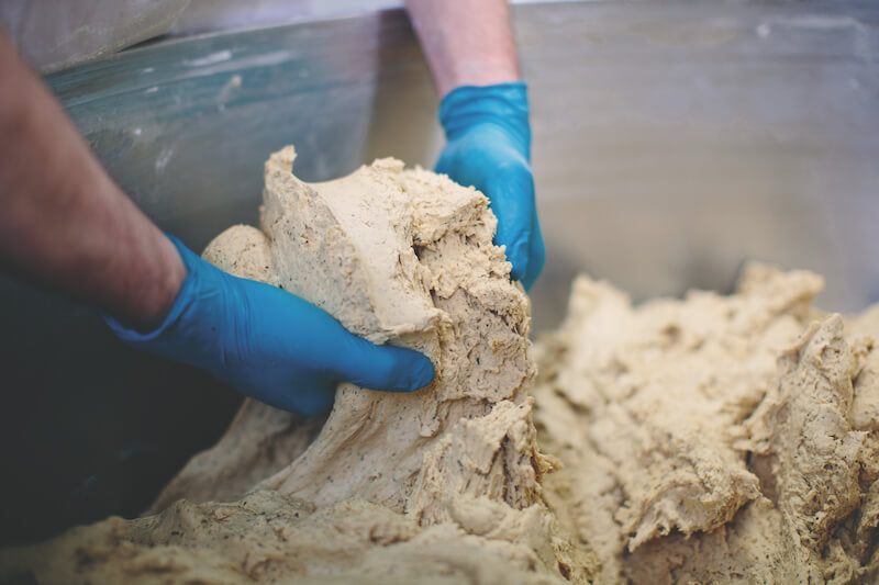 Worker wearing blue gloves folding sourdough cracker dough in a stainless-steel mixing tub at an artisanal bakery