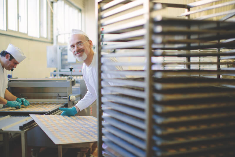 Two bakers shaping and arranging sourdough crackers on sheets in a small artisan bakery, tall drying racks in foreground