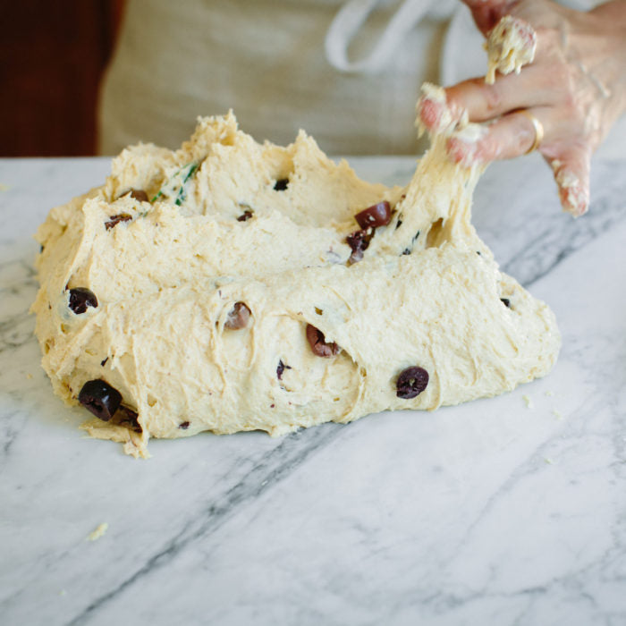 Hands folding olive-studded einkorn bread dough on a marble countertop