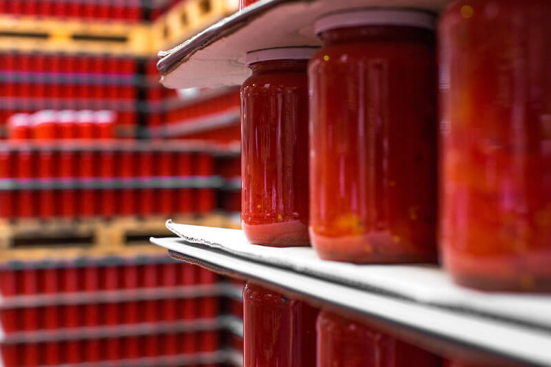 Jars of organic crushed tomatoes on a tray in a canning factory with stacked jars on shelves behind