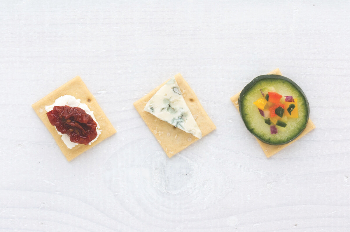Top down view of three sourdough crackers topped with sundried tomato and ricotta, blue cheese and cucumber on white wood
