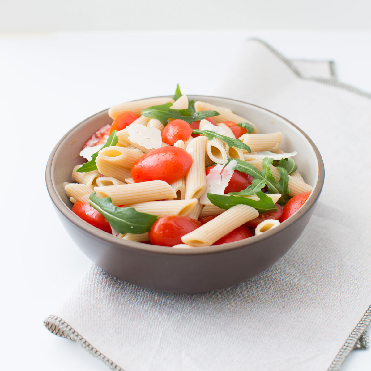 Cooked Organic Einkorn penne served in a bowl with cherry tomatoes and basil on a light napkin