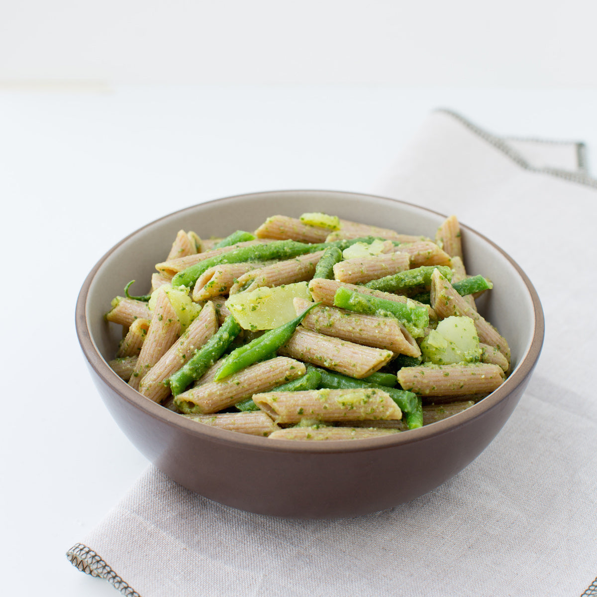 Bowl of cooked Organic Einkorn whole wheat penne with green vegetables in a brown bowl on a light cloth background