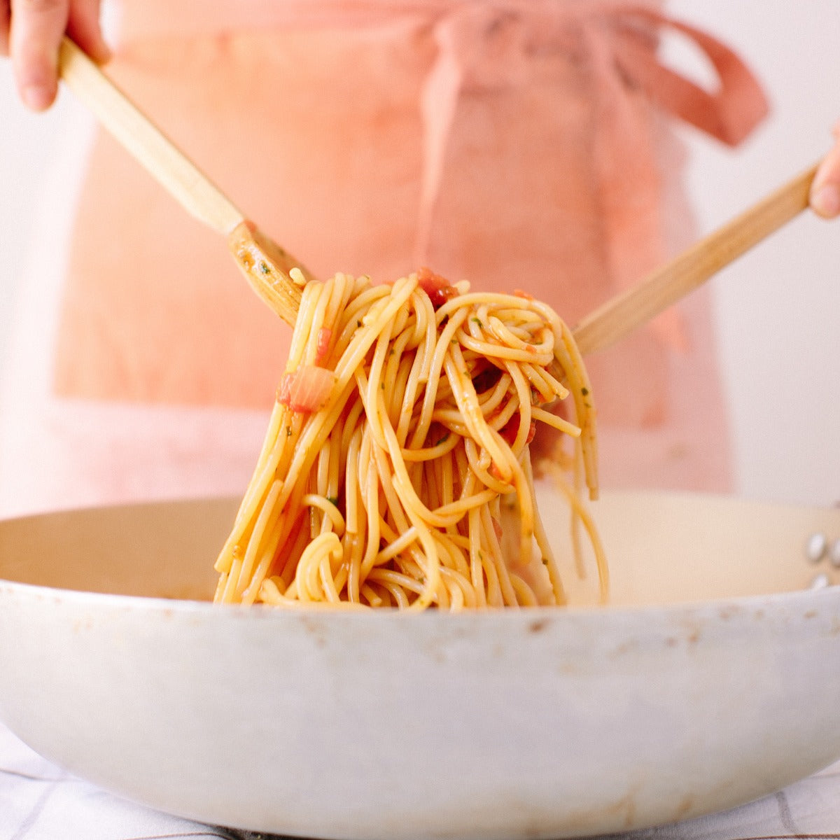 Person tossing organic Einkorn spaghetti with wooden utensils over a light skillet, pink apron in background