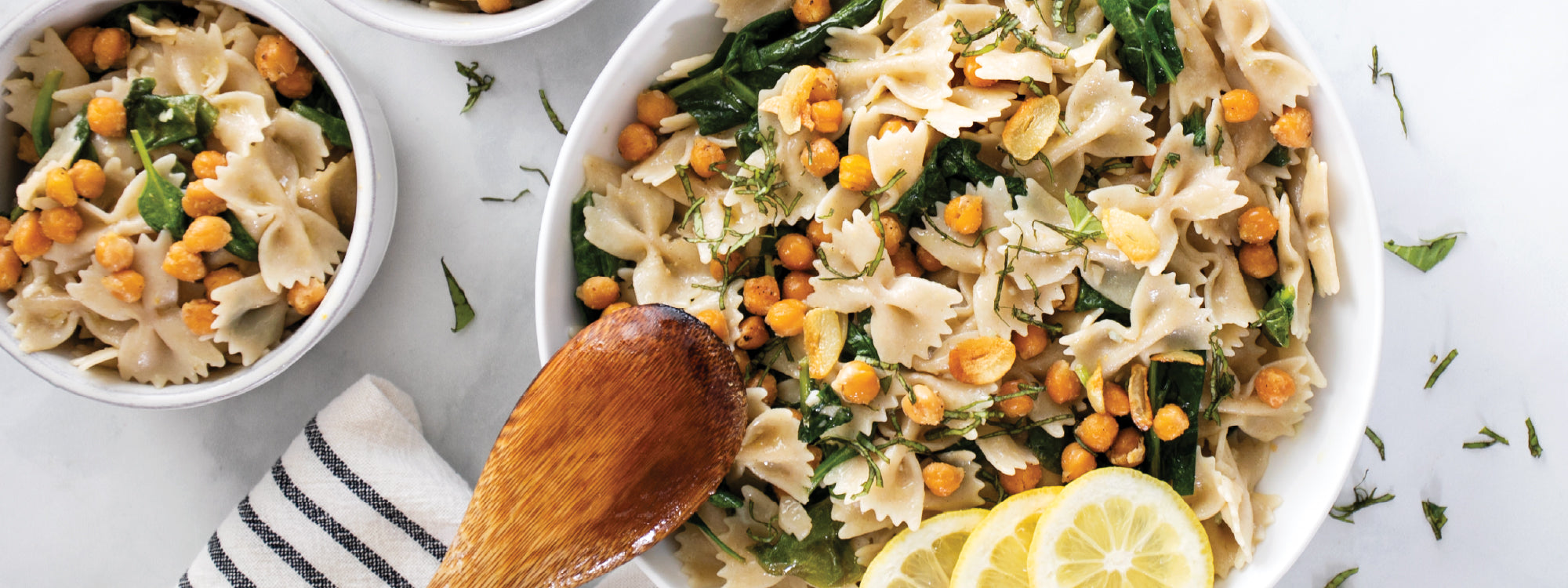 Bowl of bow-tie pasta with roasted chickpeas, spinach, lemon slices and wooden spoon on marble surface