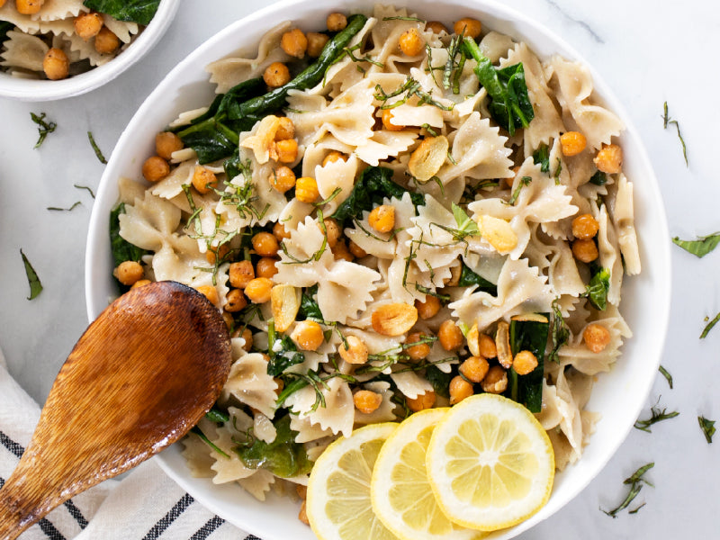 Bowl of farfalle pasta with spinach, roasted chickpeas, toasted garlic, sliced lemons and fresh basil; wooden spoon