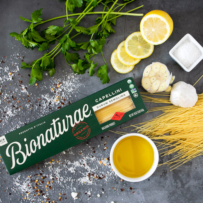 Angled view of Bionaturae Capellini box with organic durum semolina and ingredients: olive oil, garlic, lemon, parsley on dark surface