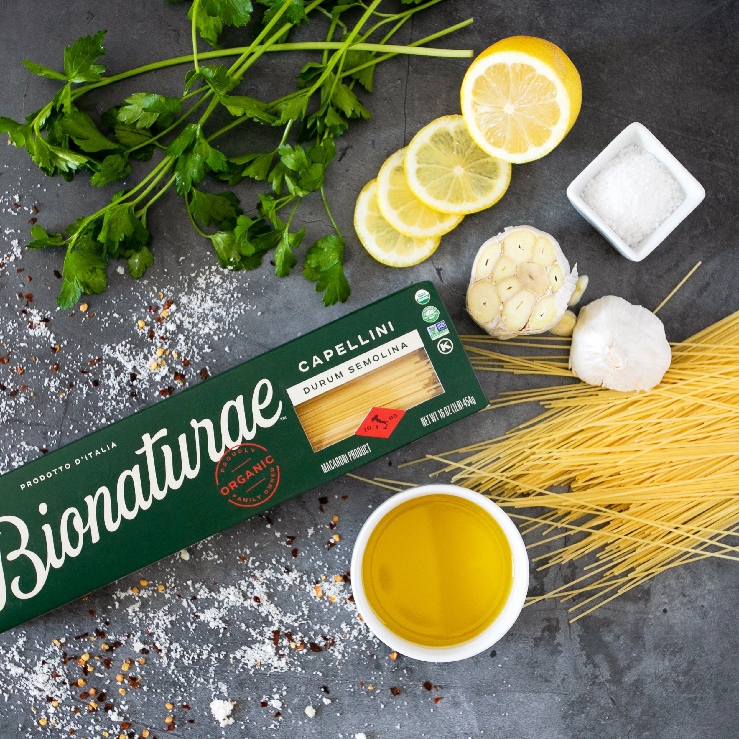 Front view of Organic Durum Semolina Capellini box showing loose capellini pasta and olive oil on a dark textured background