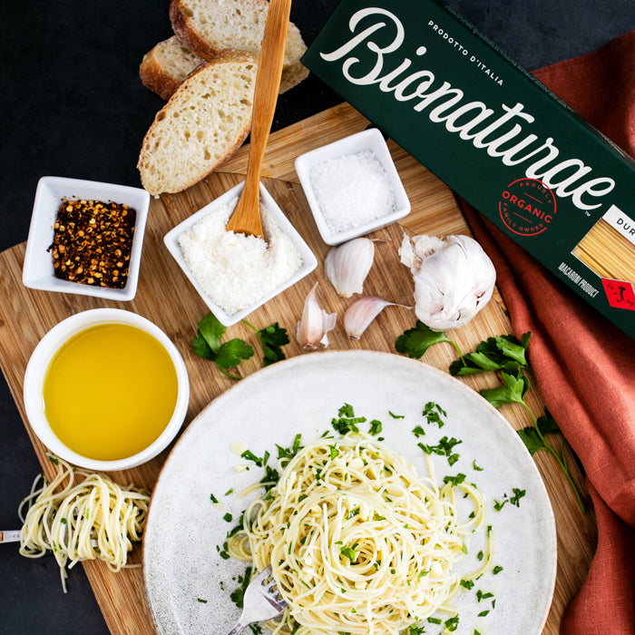 Capellini aglio e olio on a plate, with olive oil, garlic, red pepper flakes, grated cheese, bread, and Bionaturae pasta box