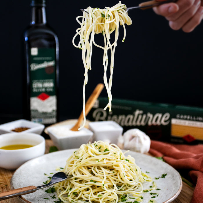 Capellini twirled on a fork above a plate of garlic and olive oil pasta, Bionaturae olive oil bottle and box in the background