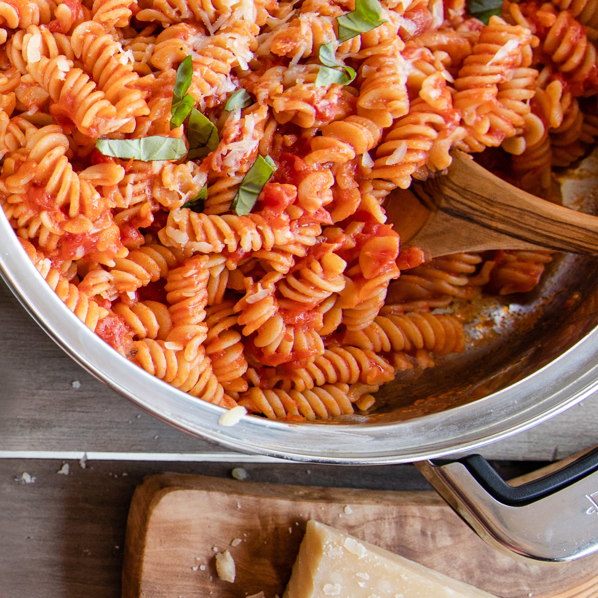 Cooked organic gluten-free rice and lentil fusilli tossed in tomato sauce with basil in a stainless-steel skillet and wooden spoon