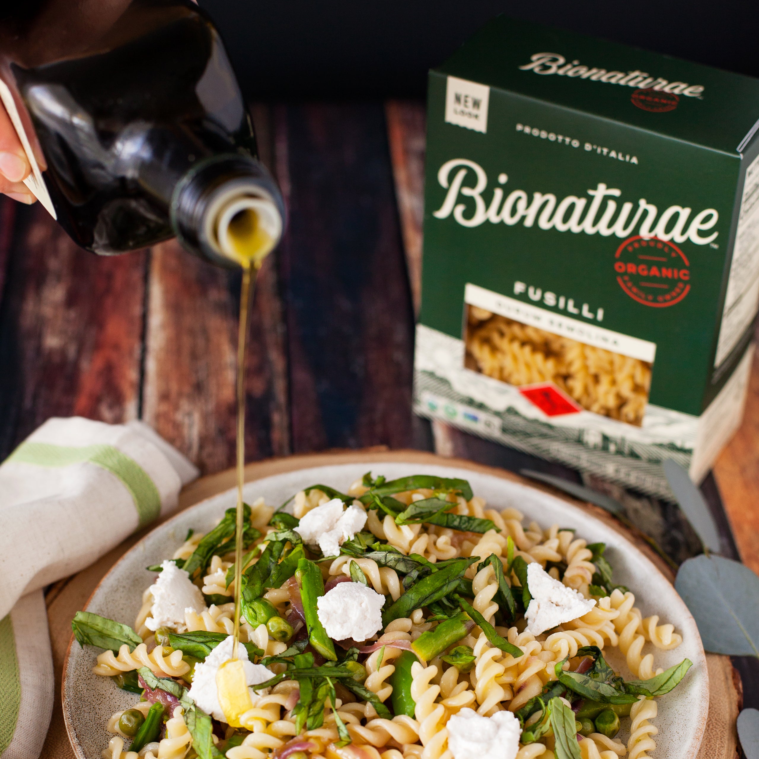 Served fusilli pasta with greens and ricotta in a skillet beside a Bionaturae pasta box and olive oil being poured