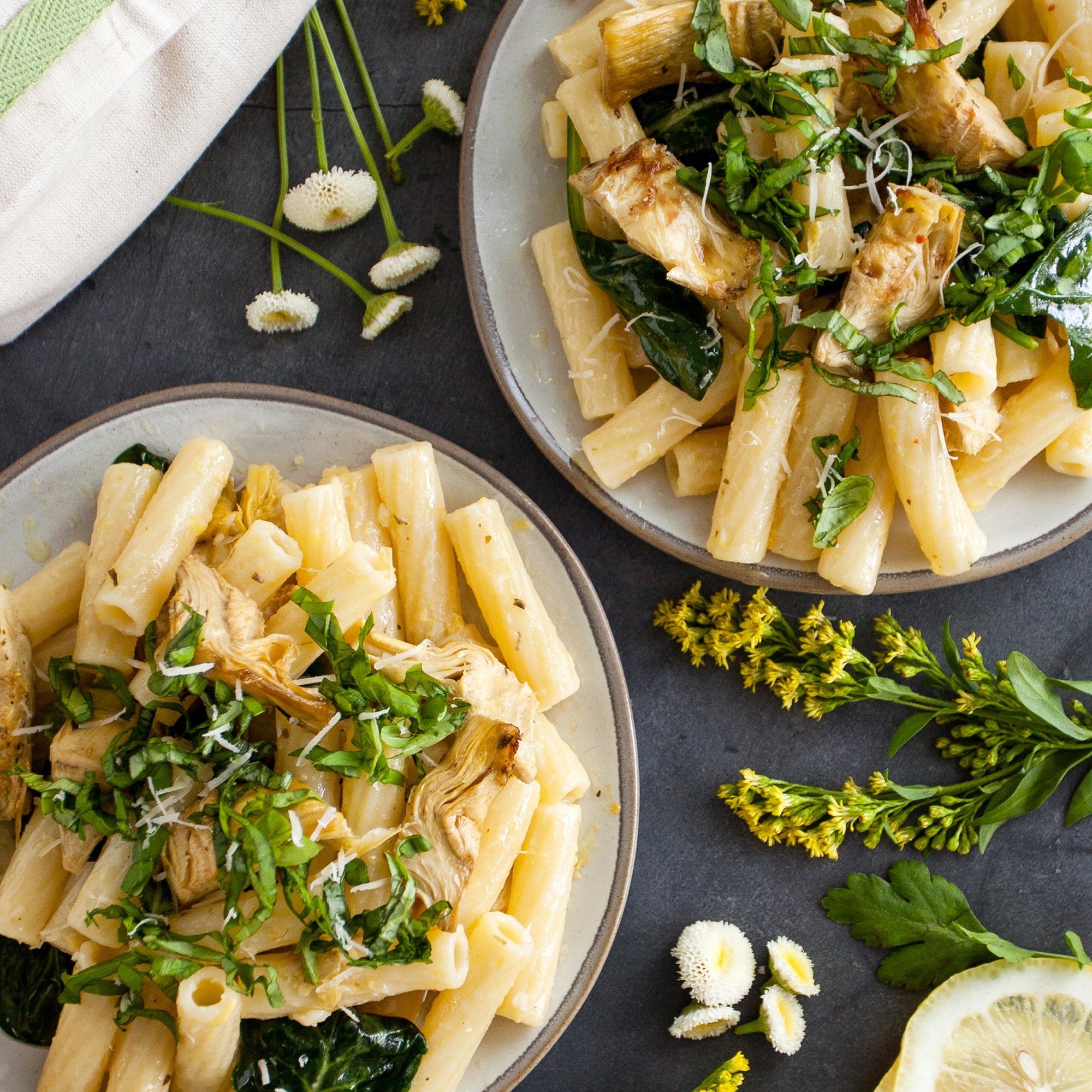 Two plates of cooked organic durum semolina rigatoni with artichokes, wilted greens and grated cheese on a dark table