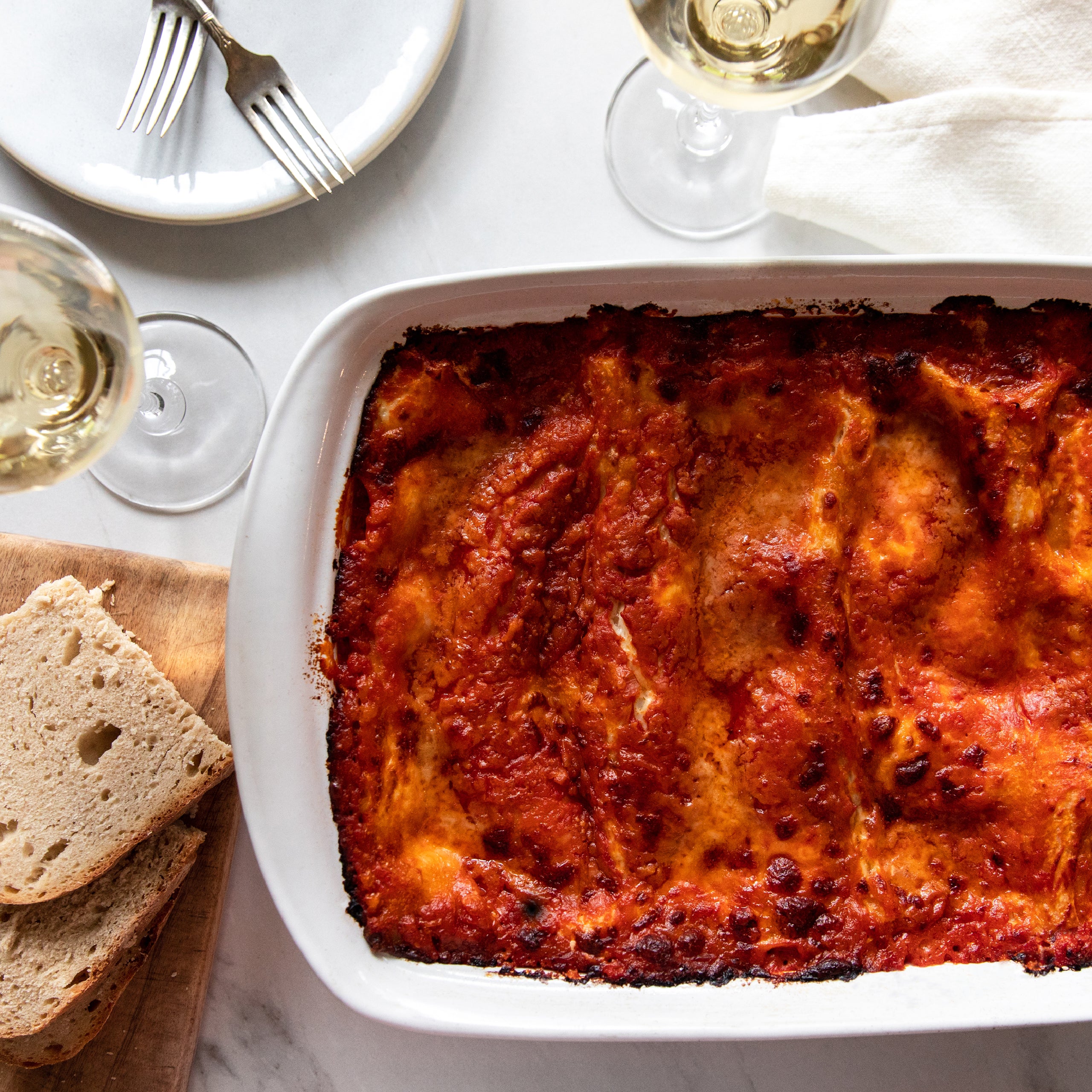 Baked organic durum semolina lasagne in a white baking dish on a table with sliced bread and a glass of white wine