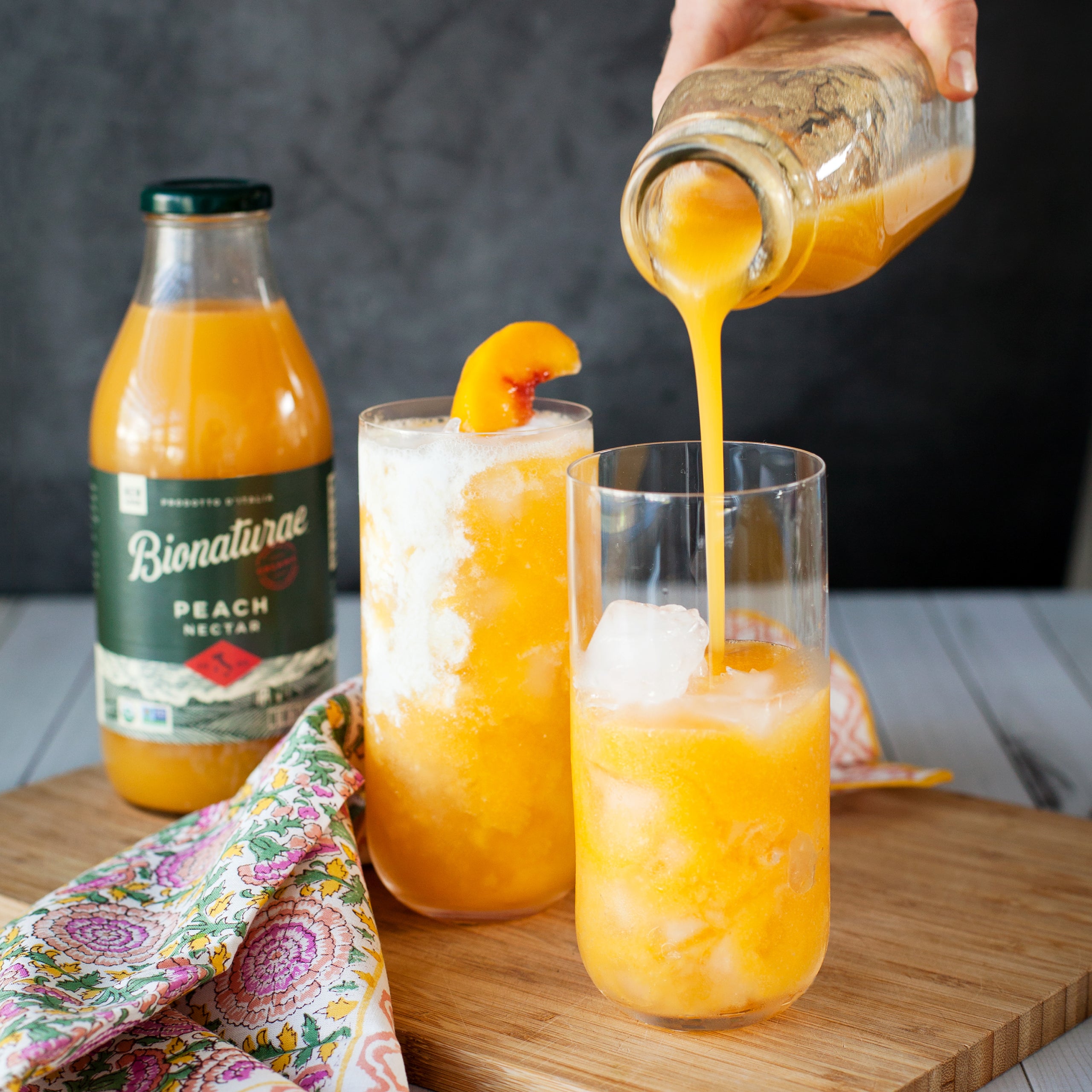 Front view of Organic Peach Nectar glass bottle showing the amber orange juice and label with a poured glass on a wooden board