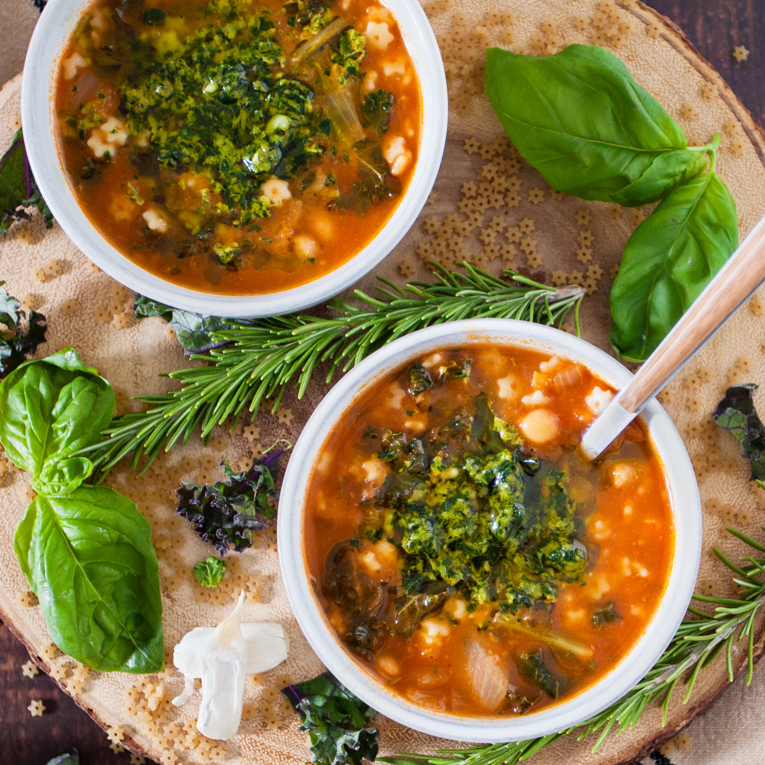 Two bowls of minestrone soup made with organic strained tomatoes, garnished with basil and rosemary on a wooden board