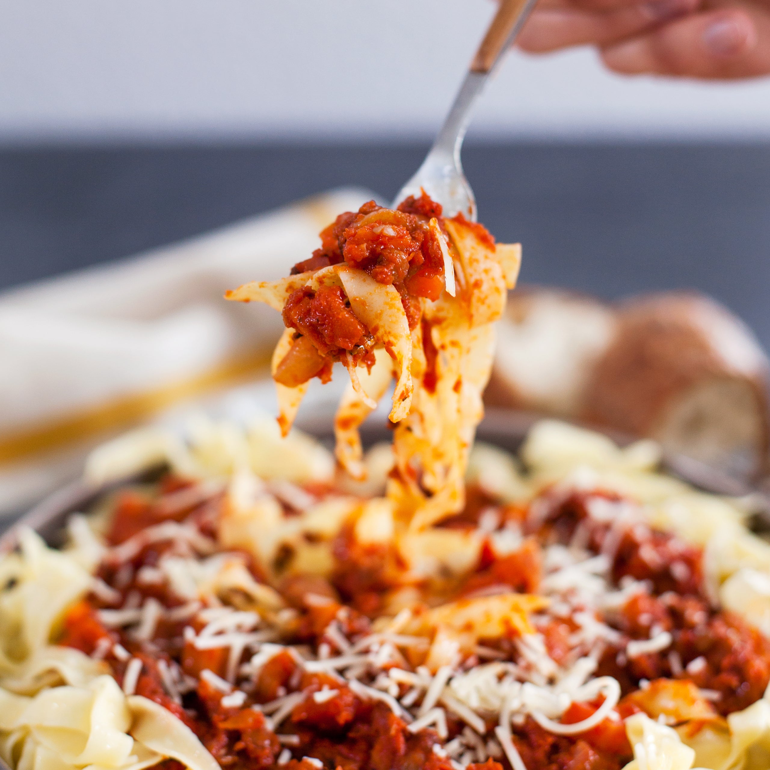 Close-up of fork lifting ribbon pasta coated in chunky organic tomato Bolognese sauce with grated cheese over a bowl