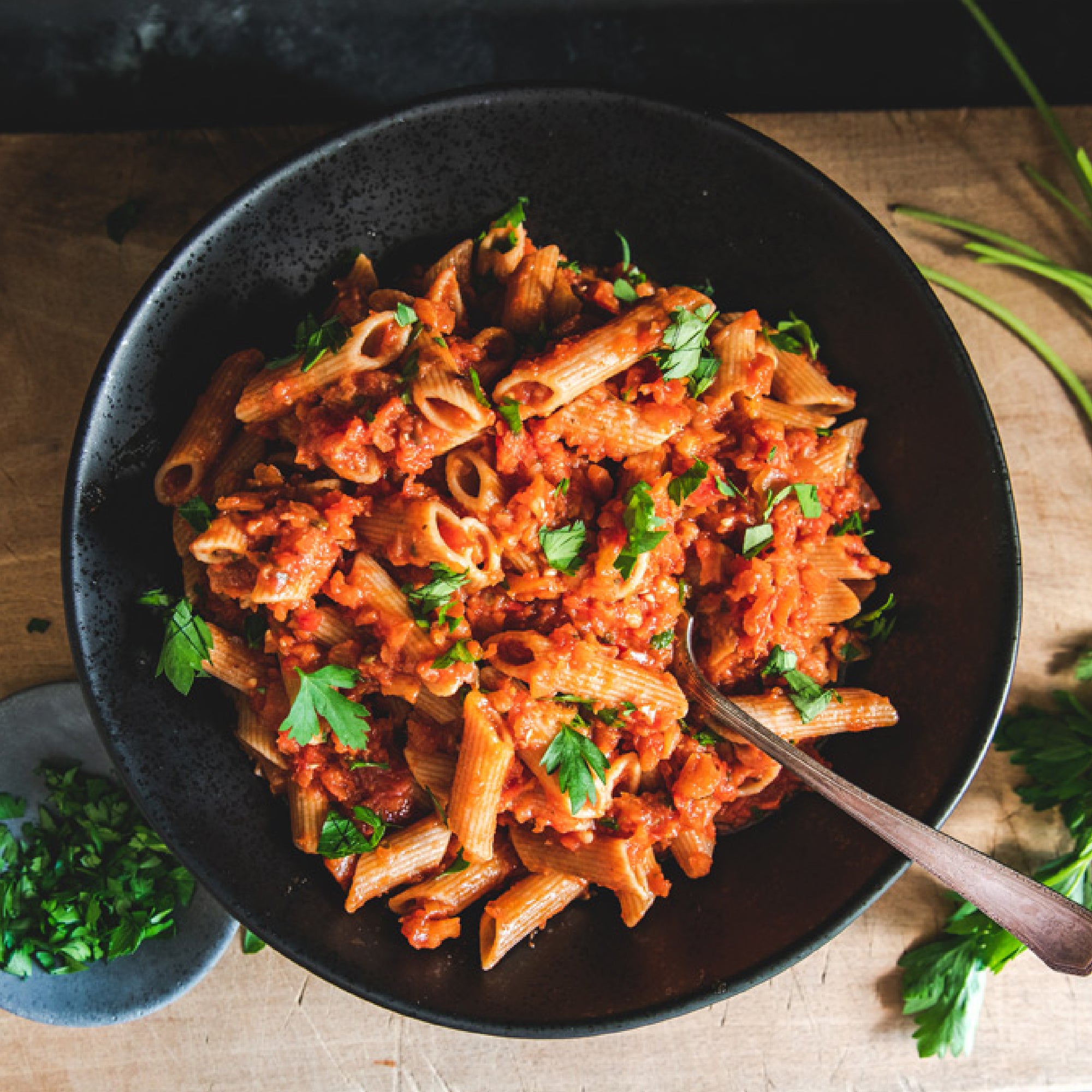 Top-down view of penne pasta tossed in chunky organic tomato sauce, garnished with fresh parsley in a black bowl