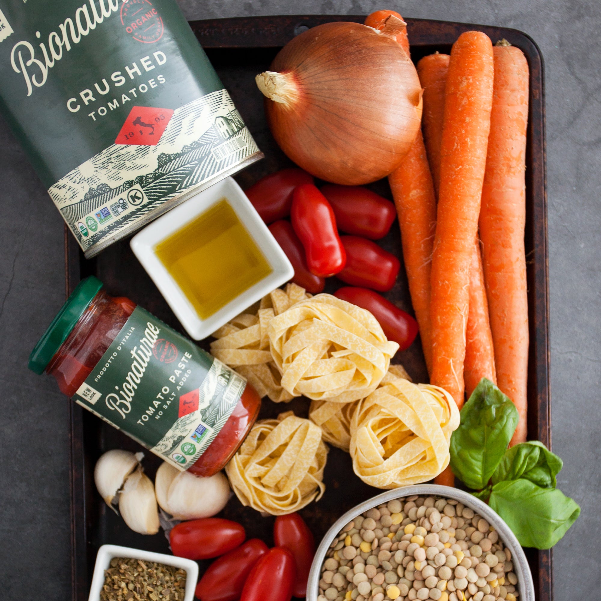 Jar and can of Bionaturae organic tomato products with pasta, garlic, carrots, basil, and olive oil on a dark tray