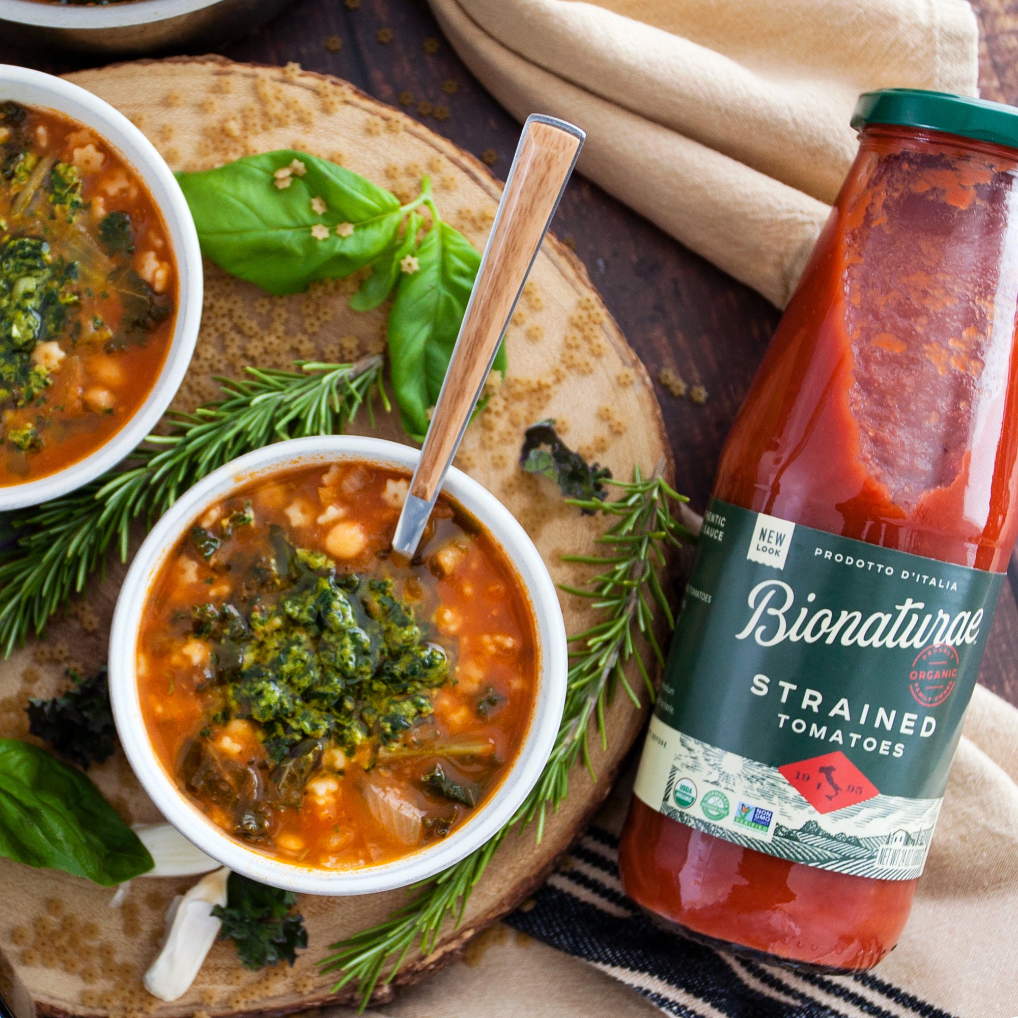 Bionaturae jar of strained tomatoes beside two bowls of tomato soup garnished with basil, pesto, and rosemary on a wooden board