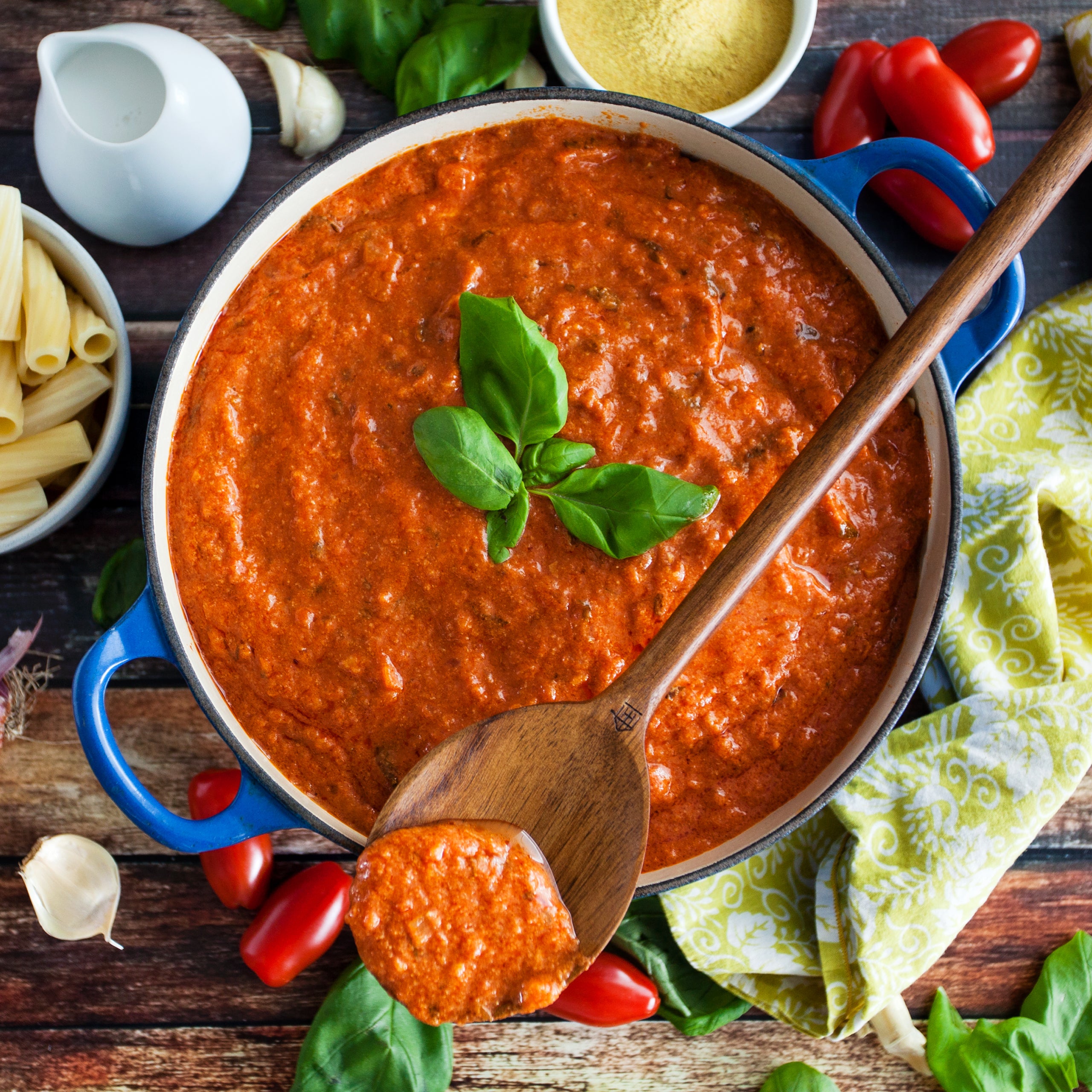 Blue enameled pot of tomato sauce with wooden spoon and basil, styled on a rustic table with fresh tomatoes and pasta