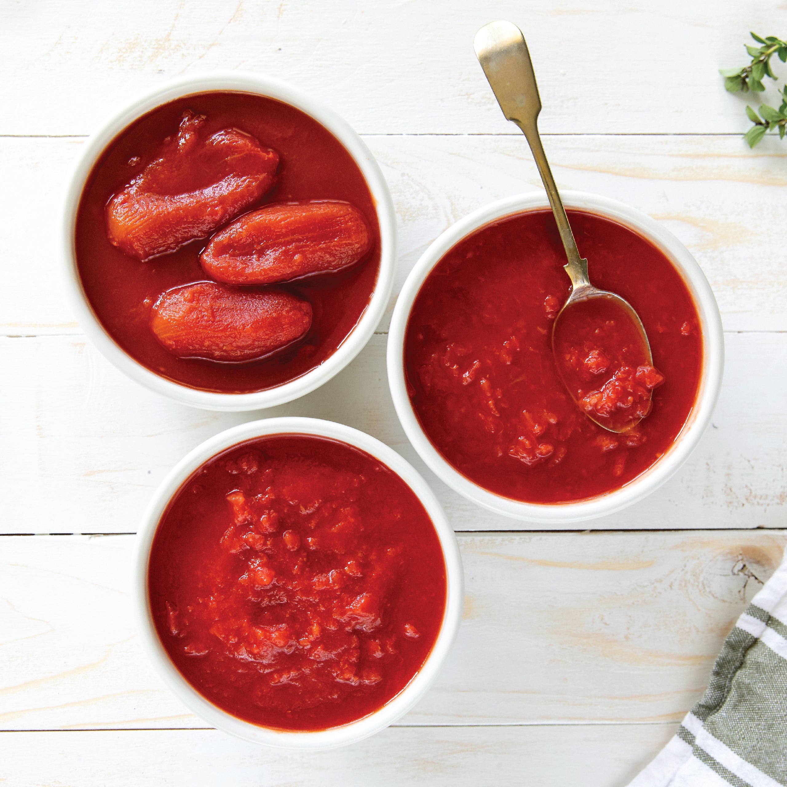 Three bowls holding whole peeled, crushed, and diced tomatoes on a white surface with a spoon and herbs
