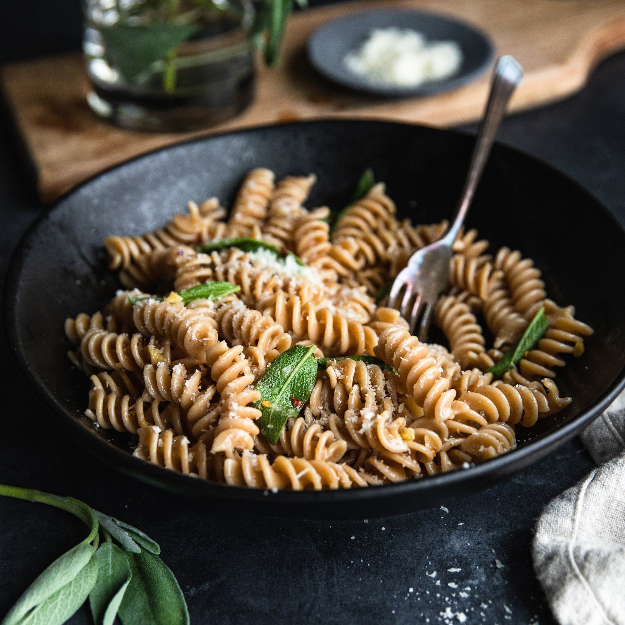 Cooked organic whole wheat fusilli in a black bowl, tossed with grated cheese and sage, fork resting in the bowl
