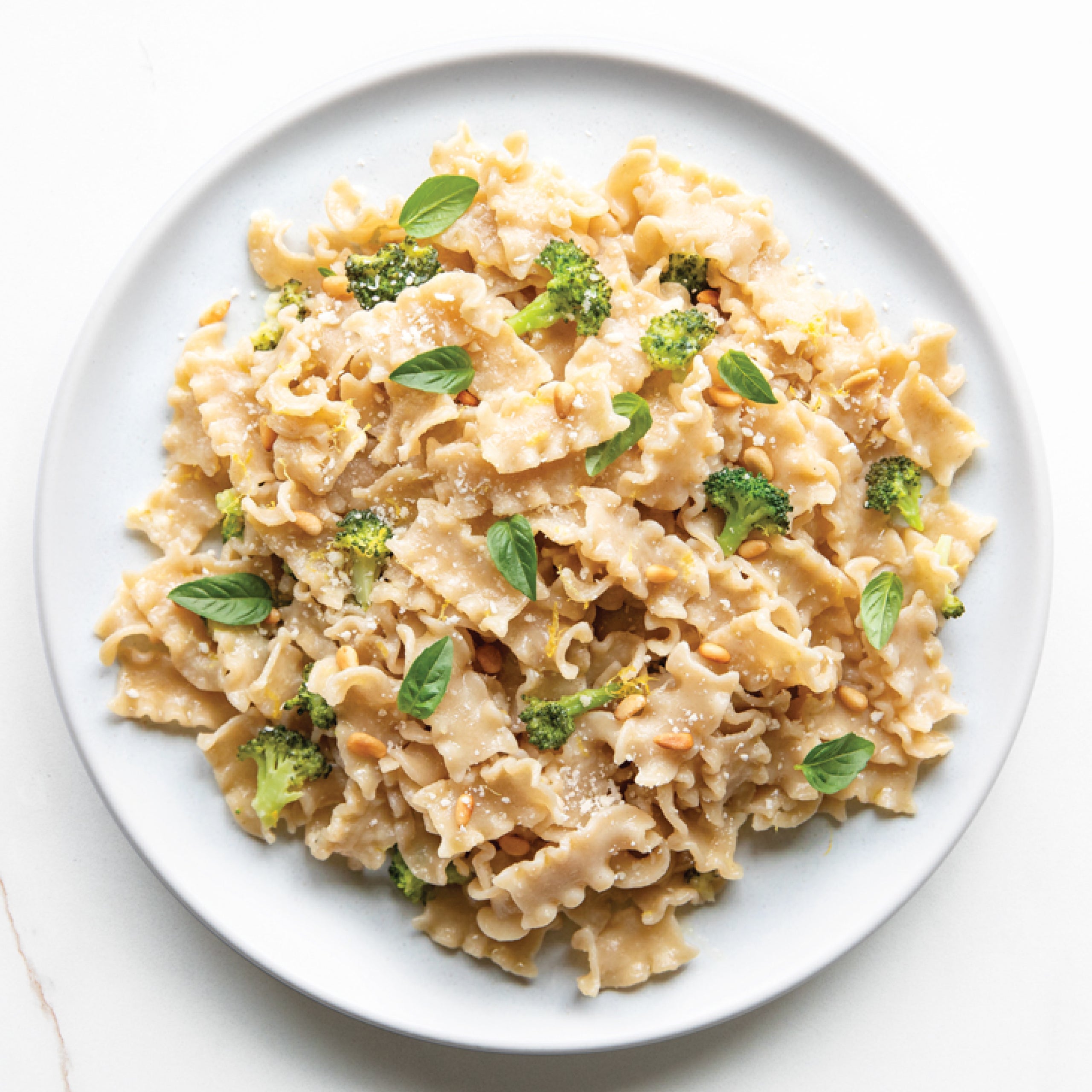 Overhead of cooked Organic Gluten Free Brown Rice Mafalda pasta with broccoli, basil, pine nuts, and grated cheese on a white plate
