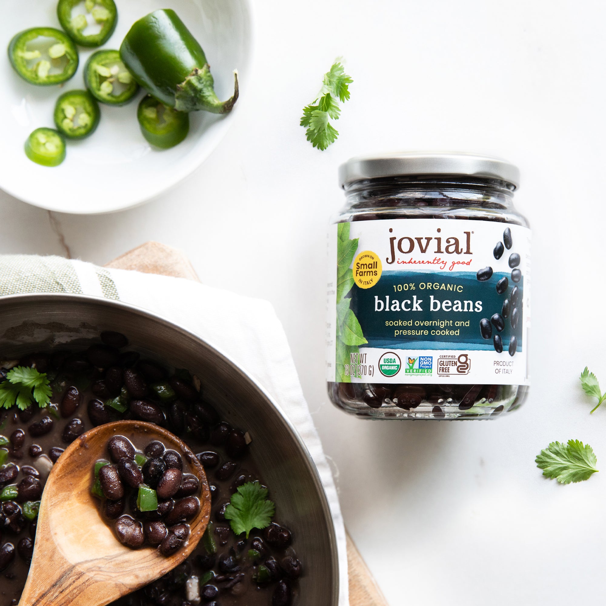 Top-down view of Jovial 100 percent Organic Black Beans jar with bowl of cooked beans, spoon, jalapeño and cilantro