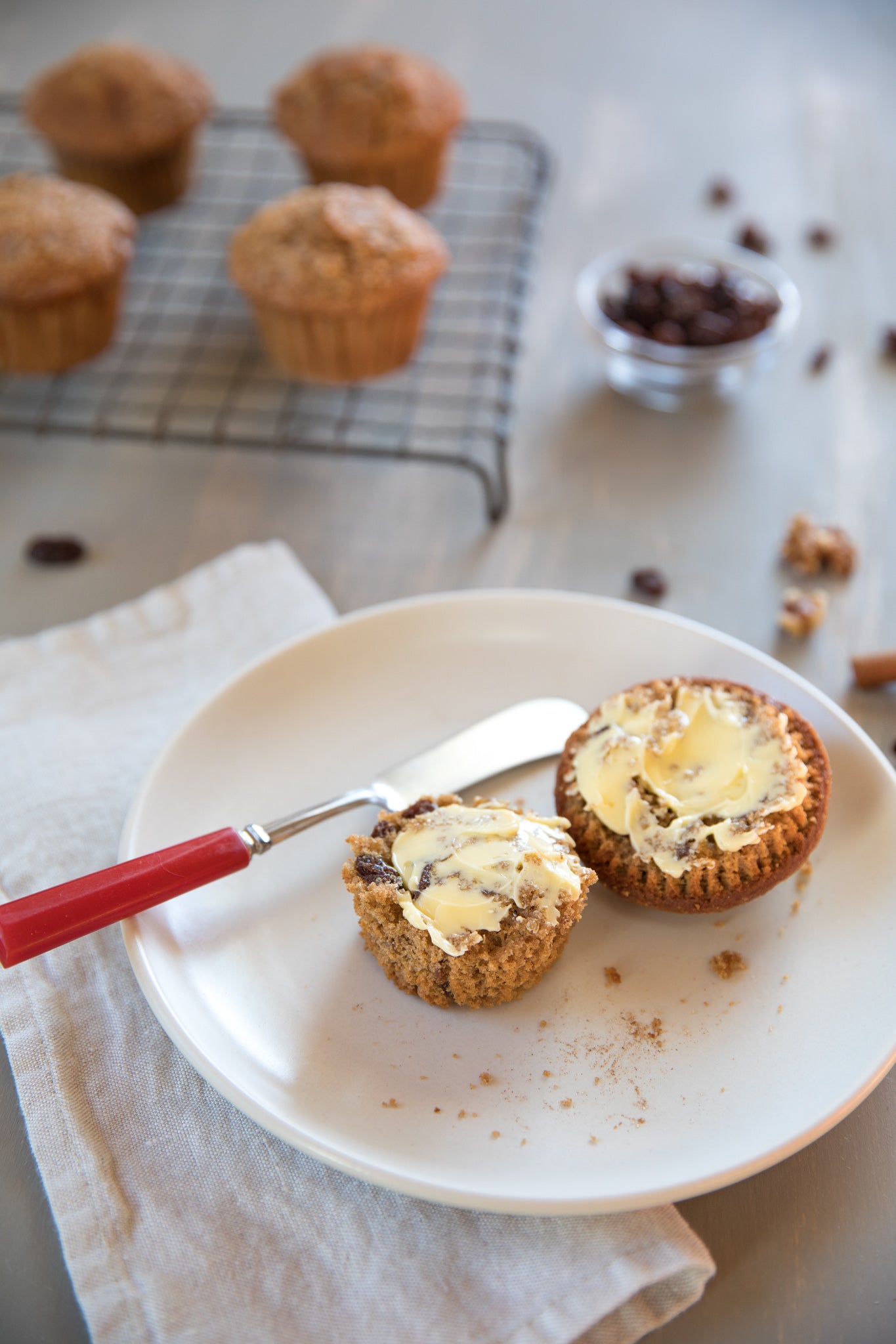 Spiced apple walnut muffins, one halved and buttered on a white plate with muffins cooling on a rack in the background