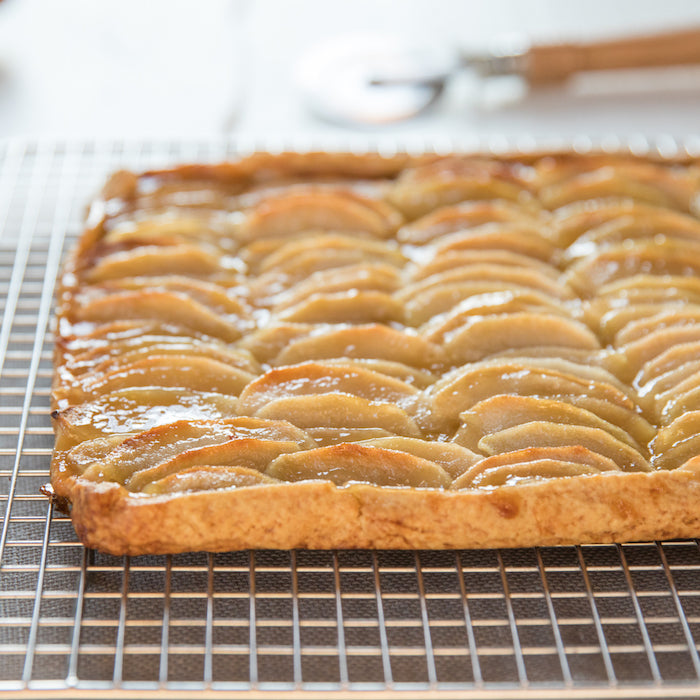 Close-up side view of an Einkorn apple caramel tart showing overlapping glazed apple slices on a golden flaky crust on a wire cooling rack