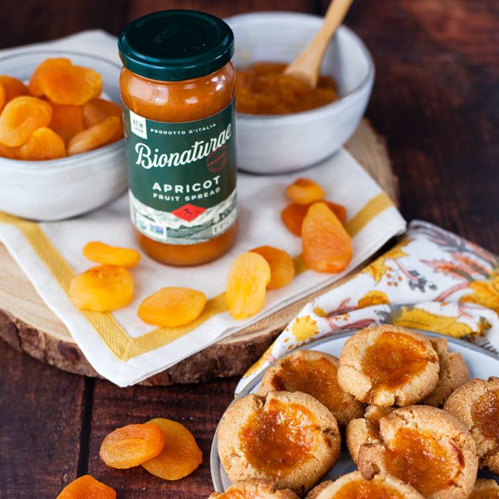 Jar of Bionaturae apricot spread beside bowls of dried apricots and a plate of apricot thumbprint cookies on a wooden table