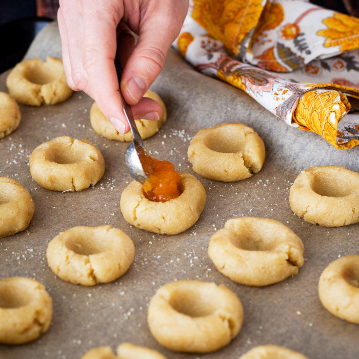 Hand spooning apricot spread into thumbprint cookie dough on a parchment-lined baking sheet with a floral napkin