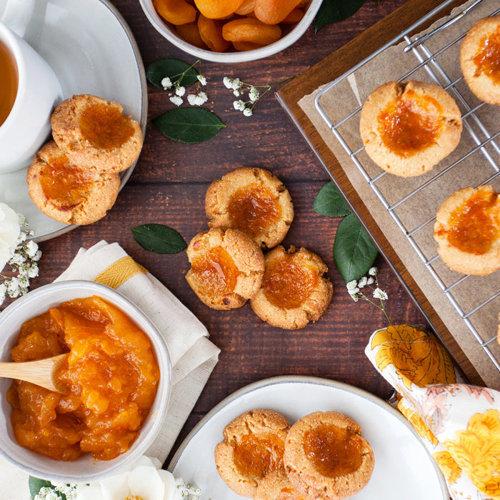Apricot thumbprint cookies on a cooling rack and plates with a bowl of apricot spread on a wooden table
