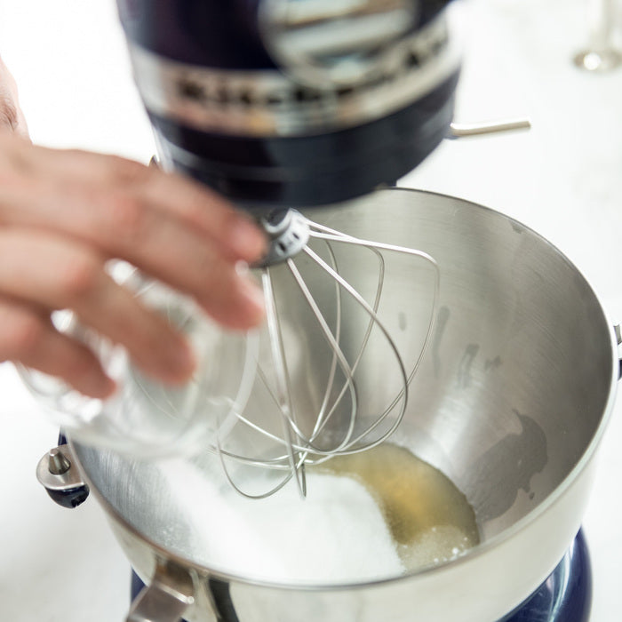 Hand pouring aquafaba into a stand mixer bowl with sugar and a whisk attachment, preparing vegan macaroon batter