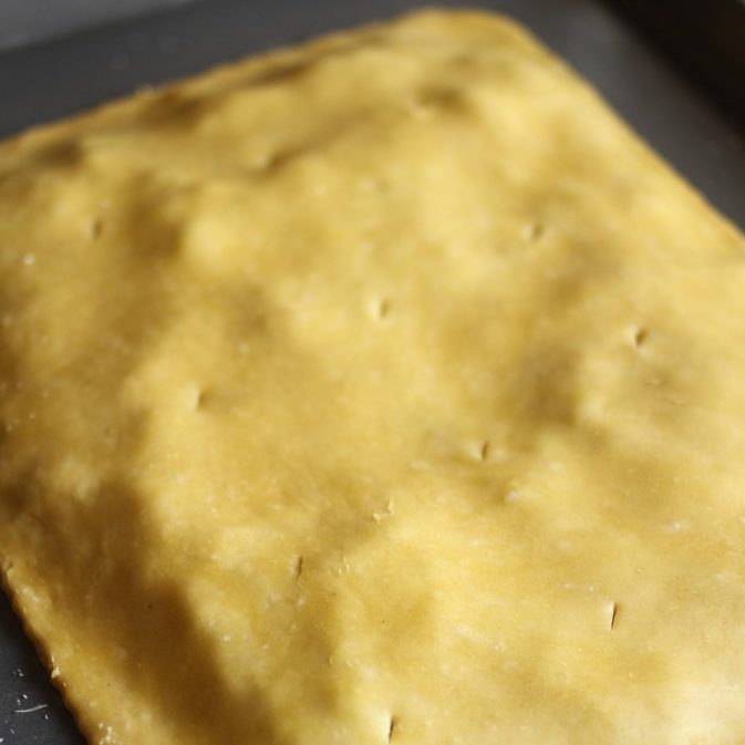 Top-down view of mixed greens pie with einkorn crust showing unbaked golden dough dotted with fork pricks on a dark baking sheet
