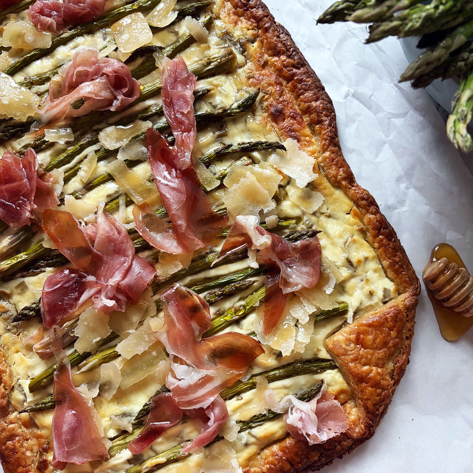 Close-up of asparagus spring tart with sourdough crust showing roasted asparagus spears, shaved Parmesan, and prosciutto on parchment